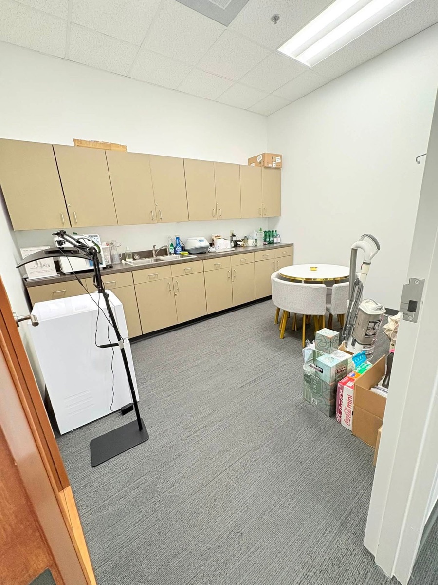 An office break room with tan cabinets, a sink counter, a round table and gray carpet